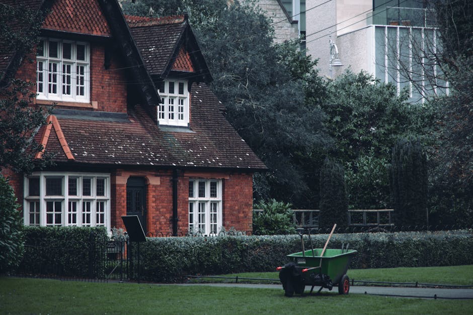 A gardener, dressed in a dark blue uniform with reflective yellow stripes, is trimming a dense, well-maintained hedge using edging shears with orange handles. The hedge features small, glossy, dark gr